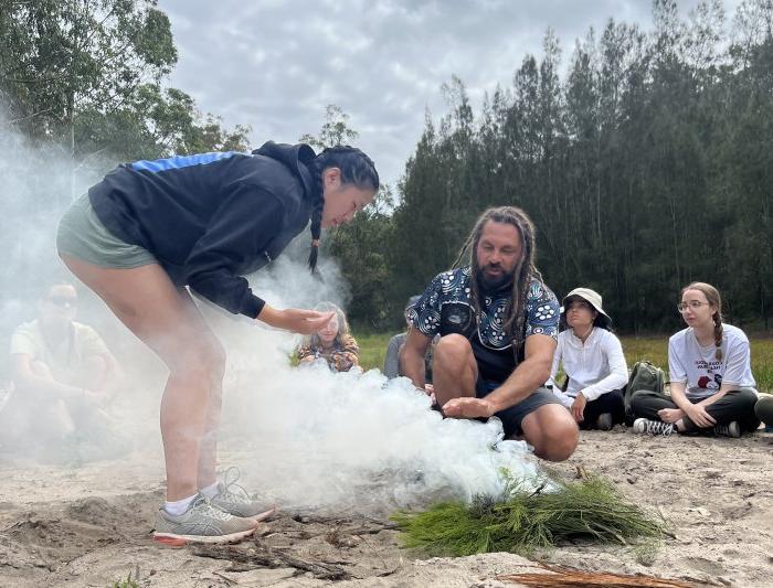 A student leans down over smoking grasses in an aboriginal custom of burning native plants to ward off bad spirits.